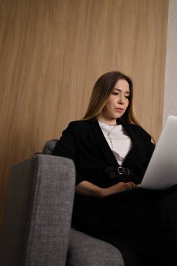 Genç bir kadın, şık bir koltukta rahat bir şekilde oturan dizüstü bilgisayarına odaklanmış. Şık iç mekan doğal ahşap zemin ile güçlendirilerek sakin bir çalışma alanı ortamı yaratıyor..