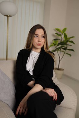 Genç bir kadın şık bir oturma odasındaki kanepede zarifçe oturur. Etrafı bitkiler ve yumuşak güneş ışığıyla çevriliyken düşünceli bir şekilde bakıyor ve sakin bir atmosfer yaratıyor..