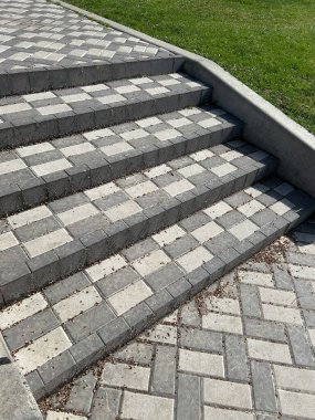 Birbirine kenetlenmiş gri ve beyaz patikalardan oluşan iyi yapılandırılmış adımlar açık mavi gökyüzünün altındaki canlı yeşil çimlere doğru yükselir. Tasarım, modern peyzajı vurgulayan geometrik bir desen sergiliyor..