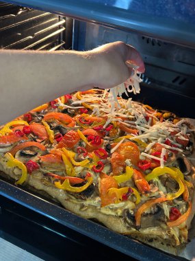 Ev fırınında yeni pişmiş sebzeli pizzanın üzerine peynir serpiştiren bir el. Renkli malzemeler arasında biber, mantar ve otlar yer alıyor. Ağız sulandıran bir tabağı servis etmeye hazır..