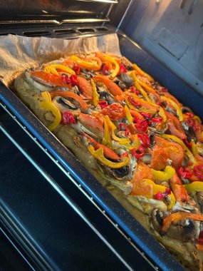 Dilimlenmiş dolmalık biber, mantar ve doğranmış domatesle kaplı bir fırında ekmek pişiriliyor. Canlı renkler iştah açıcı bir görsel yaratıyor, sağlıklı bir yemek için mükemmel..