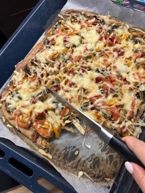 Eritilmiş peynirle kaplı lezzetli bir sebze pizzası fırın tepsisine konur. Bıçağı tutan bir el, pizzayı dilimlemeye hazır sıcak, davetkar bir mutfak atmosferinde..