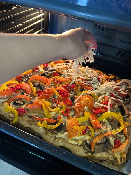 Ev fırınında yeni pişmiş sebzeli pizzanın üzerine peynir serpiştiren bir el. Renkli malzemeler arasında biber, mantar ve otlar yer alıyor. Ağız sulandıran bir tabağı servis etmeye hazır..