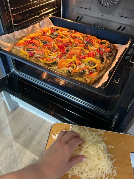 Canlı bir sebze pizzası fırında pişiyor, renkli malzemeler sergiliyor. Ön planda, peynir kesim tahtasına rendeleniyor ve yemek hazırlığına ekleniyor..