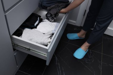 Bir insan şık bir yatak odasında açık bir çekmecede giysileri ayırıyor. Çekmecede katlanmış giysiler var, açık ve düzenli bir alan vurgulanıyor. Rahatlamış atmosfer kişisel bir dokunuşu yakalar..