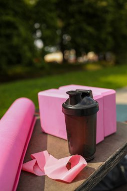 Pembe bir yoga minderi, sallanan bir şişe ve yeşil bir parktaki bantlara yerleştirilen direnç bantları güneşli bir öğleden sonra idman yaptığını gösteriyor..