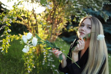Genç bir kadın yemyeşil bir bahçede elinde narin beyaz çiçeklerle oturuyor. Güneş ışığı ağaçların arasından süzülüp, sıcak bir parıltı yaratır. O, düşünceli bir şekilde güzel çevreye bakar..