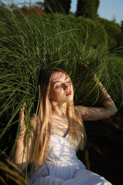 Uzun saçlı genç bir kadın yemyeşil çimenlerin arasında yavaşça oturur, sıcak güneş ışığında sırılsıklam olur. Onun barışçıl ifadesi öğleden sonra boyunca güzel bir bahçede sükuneti yansıtır..