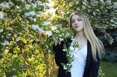 Genç bir kadın çiçek açan bitkilerle çevrili verimli bir bahçede zarif bir şekilde duruyor. Sıcak güneş ışığı huzurlu bir atmosfer yaratıyor, sakin ifadesini arttırıyor..