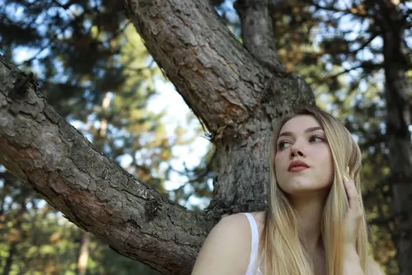 Uzun, dalgalı saçlı genç bir kadın, sessiz bir ormanda yemyeşil yaprakların arasından süzülen güneş ışığıyla çevrili, dokulu bir ağaç gövdesine karşı huzur içinde oturur..