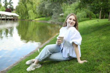 Genç bir kadın dingin bir gölün yakınındaki yumuşak çimenlerde oturuyor, sıcak bir içecek tutuyor ve gülümsüyor. Yerleşim verimli yeşillik ve hafif dalgalı su içeriyor. Huzurlu bir atmosfer yaratıyor..