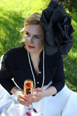 Siyah bluzlu ve beyaz etekli bir kadın yeşil çimlerde rahatlıyor, bardaktan bir içecek yudumluyor. Büyük siyah bir çiçek, bahçede huzurlu bir öğleden sonranın tadını çıkarırken şapkasını süslüyor..