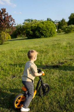 Genç bir çocuk yemyeşil bir parkta parlak turuncu bir denge bisikleti sürüyor. Anne, güneşli bir öğleden sonra rehberlik ve cesaret vermek için yakınlarda duruyor. Ağaçlar ve çalılar etraflarını sarmış..