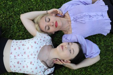 Parktaki yeşil çimlere uzanmış güneşli bir günün tadını çıkaran iki genç kadın. Barışçıl ifadeleri ve rahat vücut dilleri var, sakin bir açık hava ortamı ile çevrili..