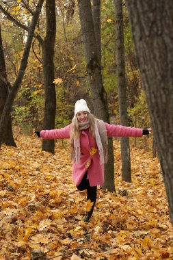 Canlı bir sonbahar ormanında, pembe paltolu ve beyaz şapkalı bir kadın düşmüş yaprakların arasında neşeyle kıvrılır. Altın yapraklar onu çevreleyip mevsimsel zevklerin resimsel bir sahnesini oluşturuyor..