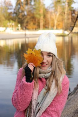 Sonbahar boyunca sakin bir gölün kenarında duran bir kadın, elinde parlak altın yapraklar ve gülümsüyor. O rahat beyaz bir şapka takıyor ve sıcak bir eşarba sarılmış. Arka plandaki ağaçlar sonbahar renklerini gösterir.