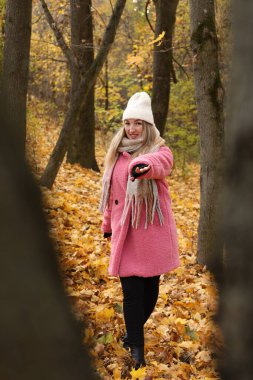 Pembe ceketli ve beyaz şapkalı bir kadın ağaçların arasında duruyor. Ormanda sarı ve turuncu yapraklardan oluşan bir halıdan geçerken neşeyle uzanıyor..