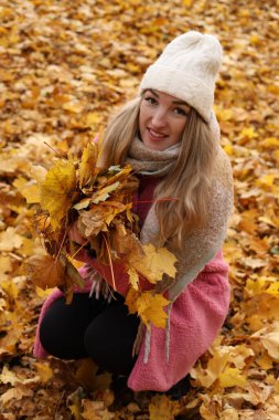 Canlı sonbahar yapraklarıyla dolu bir parkta çömelmiş bir kadın bir avuç dolusu sarı ve turuncu yaprak topluyor. Güneş onun rahat elbisesinin üzerinde ışıldıyor..
