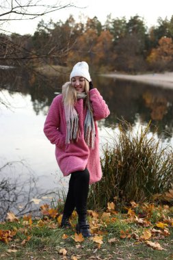 Huzurlu bir gölün yanında duran genç bir kadın rahat pembe bir ceket ve sıcak aksesuarlar giyiyor. Parlak sonbahar yaprakları gülümserken resim gibi bir zemin oluşturuyor..