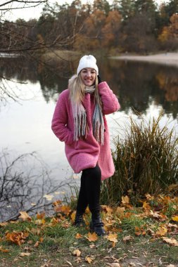 Bir sonbahar ortamında, bir kadın sakin bir gölün yanında duruyor, sıcak pembe bir ceket ve beyaz bir şapka giyerek gülümsüyor. Etrafına saçılmış yapraklar, doğanın resimli bir sahnesini oluşturuyor..