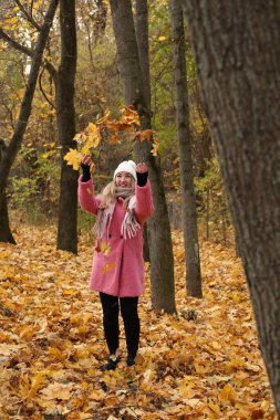 Pembe paltolu ve beyaz şapkalı bir kadın ormanda duruyor, neşeyle sonbahar yapraklarını havaya savuruyor. Etrafındaki ağaçlar canlı sonbahar renkleri sergiliyor, neşeli bir atmosfer yaratıyor..
