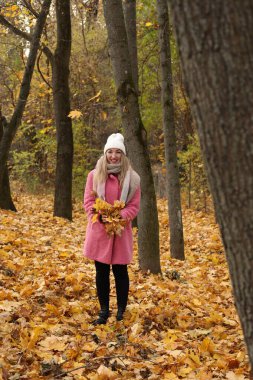 Genç bir kadın, elinde bir buket sarı ve turuncu yaprakla, sonbahar manzarasının tadını çıkarırken, pembe bir ceket ve beyaz bir şapka giyerek ormanda canlı yaprakların arasında duruyor..
