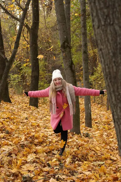 Canlı bir sonbahar ormanında, pembe paltolu ve beyaz şapkalı bir kadın düşmüş yaprakların arasında neşeyle kıvrılır. Altın yapraklar onu çevreleyip mevsimsel zevklerin resimsel bir sahnesini oluşturuyor..