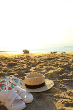 Gün batımında kumlu bir plajda, renkli sandaletler ve engebeli arazide dinlenen hasır bir şapkayla dinlenmek. Güneş ufukta sıcak bir parıltı yayıyor..