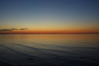 Güzel bir günbatımı, plajın sakin suları üzerinde sergilenir. Sıcak turuncu ve mavi renkler gökyüzüne saçılır. Yumuşak dalgalar kıyıya vurur, huzurlu ortamı güçlendirir..