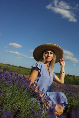 Lavanta tarlaları, mavi elbiseli bir kadının manzaranın tadını çıkardığı mesafeye kadar uzanıyor. Etrafı canlı çiçeklerle çevriliyken geniş kenarlı bir şapka takıyor ve gülümsüyor..