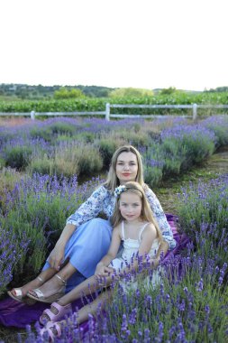 Bir kadın ve kızı, çiçek açan lavantanın ortasında, huzurlu ortamın tadını çıkarıyorlar. Her ikisi de açık renk giyinmiş, kameraya dönük gülümsüyor, mor çiçeklerle çevrili..