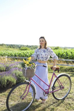 Genç bir kadın, güzel bir lavanta tarlasında çiçeklerle süslenmiş pembe bir bisikletin yanında duruyor. Güneş ışıl ışıl parlıyor, çiçekli bluzunu ve canlı manzarasını aydınlatıyor..