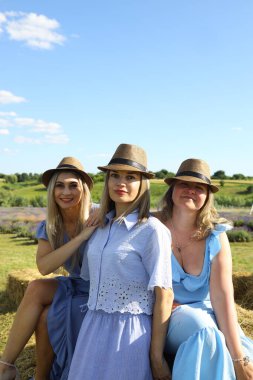 Üç arkadaş açık mavi gökyüzünün altında canlı bir lavanta tarlasında birlikte oturuyorlar. Yaz kıyafetleri ve hasır şapkalar giyiyorlar, günün sıcaklığını kucaklıyorlar ve kalıcı anılar yaratıyorlar..