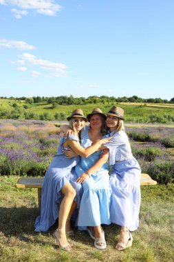 Güneşli bir yaz gününde, zarif mavi elbiseler ve şık şapkalar giyen üç kadın, çiçek açan lavanta tarlasında mutlu bir şekilde poz veriyor. Sıcak bir kucaklaşmayı paylaşıyorlar, dostluk yayıyorlar..