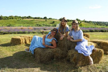 Yazlık elbiseler giymiş üç kadın açık bir gökyüzünün altındaki lavanta tarlasında saman balyalarında neşe içinde poz veriyor. Çevresindeki tepeler manzaralı kırsal alanı tamamlıyor..