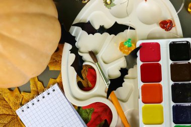 Bir masa, içinde boyanmış bir balkabağı, yarasalar, canlı renklere sahip bir palet ve dağınık yaprakların da bulunduğu çeşitli Cadılar Bayramı el işi malzemelerini sergiler. Yaratıcılık havayı doldurur..