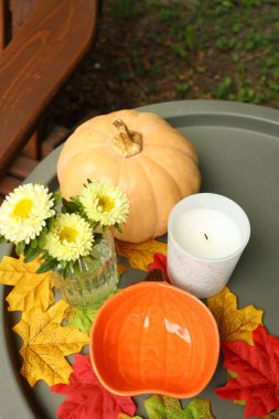 Sakin bir sonbahar masasında hafif turuncu bir balkabağı, parlak sarı çiçekli cam bir vazo ve renkli yaprakların arasında beyaz bir mum bulunur. Mevsimlik dekor için mükemmel.