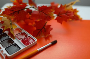 Parlak kırmızı ve turuncu yapay yapraklar, boya dolu bir paletin etrafını sarmış durumda. Canlı turuncu arka planda bir fırça duruyor. Bu rahat ortam yaratıcılığı ve sıcaklığı davet ediyor..