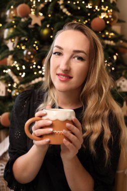 Genç bir kadın gülümserken sıcacık bir kupa tutuyor. Güzel ışıklandırılmış bir Noel ağacıyla süslenmiş, sıcak ve neşeli bir atmosfer yaratıyor..