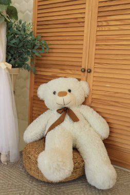 Büyük, tüylü, kahverengi kurdeleli bir oyuncak ayı ahşap bir dolabın yanındaki dokuma sepetin üzerinde rahat bir şekilde oturur ve odada sıcak ve davetkar bir atmosfer yaratır..