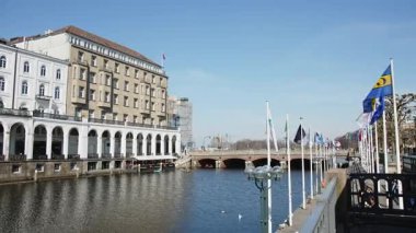 HAMBURG, GERMANY - 27 Mart 2022: Alsterarkaden, Alster Çarşısı, nehir kıyısında bir alışveriş merkezi, Canal Alster ve Jungfernstieg güneşli bir bahar gününde mavi gökyüzü