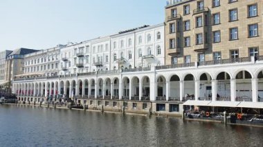 HAMBURG, ALMANY - 27 Mart 2022: Alsterarkaden, Alster Çarşısı, nehir kıyısında bir alışveriş merkezi, güneşli bir bahar gününde mavi gökyüzü ile Canal Alster. Restoranlarda.