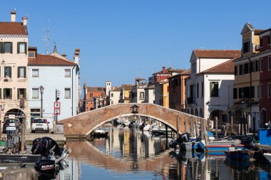 Chioggia, İtalya - 3. Mar 2025: Vena Kanalı üzerindeki tuğla köprü, tarihi merkezde demirli tekneler ve renkli binalar