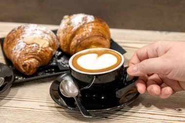 Kalp şeklinde latte sanatıyla kapuçino ve rustik bir masada taze kruvasan, fincanı tutan bir elle.