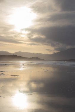 Sahilde parlak sarı günbatımı ve arka planda dağlar. Yüksek kalite fotoğraf. Büyük dalgaları olan deniz güzel bir manzara yaratıyor. Sıcak ve estetik bir atmosfer. Parlak ışık yansıma oluşturur