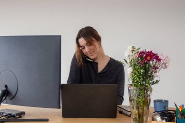 Genç Avrupalı beyaz kadın evde dizüstü bilgisayarla çalışıyor. Kaliteli fotoğraf. Bayan telefonda sohbet ediyor ve dizüstü bilgisayarından notlar alıyor. O gülümsüyor. Masa ayarlanabilir, ona izin veriyor.