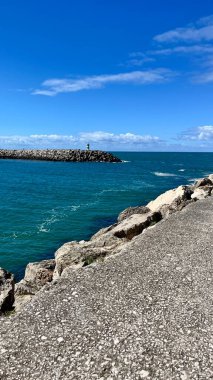 Turkuaz sular, yumuşak beyaz bulutların altında arka planda bir kıyı şeridi bulunan iskelenin kayalarına hafifçe çarpar. Dinginliğin ve doğal gücün canlı bir sahnesi..