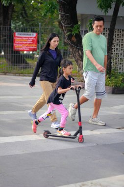 Endonezya, Surakarta - 16 Kasım 2024: Ailesinin neşe içinde yürüttüğü, sokakta scooter oynayan küçük bir çocuk