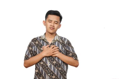 Eli göğsü ve gözleri kapalı batik tişört giyen Asyalı bir adam. Yüzünde rahatlama ve minnettarlık ifadesi var. Arkaplan düz beyaz.