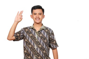 Batik elbiseli yakışıklı Asyalı adam elini izole edilmiş beyaz arka planda sallayarak insanları selamlıyor.